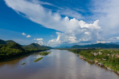 Mekong Nehri 'nin yemyeşil tepeler ve Laos' un nehir kenti Luang Prabang arasında kıvrılarak döndüğü geniş bir manzara. Yükselen kümülüs bulutları ve canlı mavi bir gökyüzü parlak gün ışığında çarpıcı bir tropikal manzara yaratır..