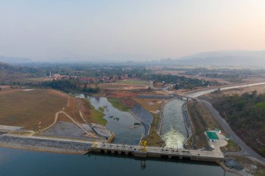 Modern bir hidroelektrik barajın havadan görüntüsü suyu düz bir kanala boşaltıyor, etrafı mühendislik arazileri, yama alanları ve puslu gün ışığı altında uzak bir köy ile çevrili. Sahne, altyapı ve açık kırsal ve alçak tepeleri birleştiriyor.