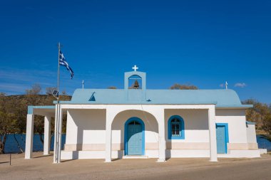 Kilada 'da berrak mavi gökyüzünün altında sakin suyun yanında kemerli girişi ve çan kulesi olan geleneksel beyaz ve mavi şapel. Güneş ışığı, minimalist cepheye taze gölgeler düşürüyor, sakin bir Akdeniz havasını çağrıştırıyor..