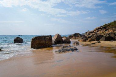 Hindistan 'ın Kovalam kentindeki Samudra Sahili' nde hafif dalgalar kıyıya vururken, büyük pürüzsüz kayalar ıslak altın kum üzerinde dinlenir. Sahne aydınlık ve huzurlu, bulutlu bir gökyüzü altında, engebeli uçurumlar uzakta..