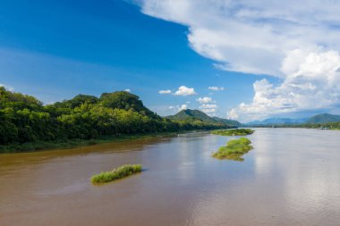 Luang Prabang, Laos yakınlarındaki yemyeşil orman bankalarını ve küçük çimenli adaları geçerek Mekong Nehri 'nin geniş manzarası. Sahnede sakin kahverengi su, canlı yeşil bitki örtüsü ve canlı mavi gökyüzünün altında dramatik kümülüs bulutları yer alıyor..