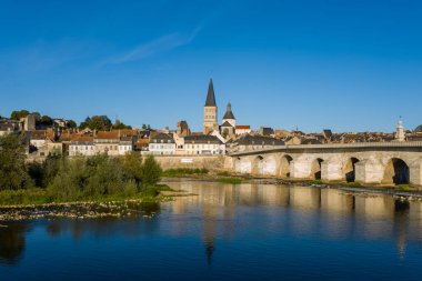 Tarihi taş köprü Loire nehri boyunca uzanır ve ortaçağ manastırına doğru ilerler ve La Charite sur Loire 'ın çatıları kümelenir. Parlak güneş ışığı ve berrak mavi gökyüzü, sakin nehir kenarı manzarasını ve geleneksel mimariyi ön plana çıkarıyor..