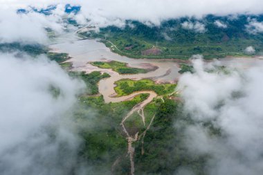 Kıvrımlı bir nehir ve çamurlu bir gölün havadan görünüşü alçak bulutlarla kısmen örtülmüş, yoğun yeşil tepeler ve ormanlarla çevrili. Manzarada doğal su kanalları, yemyeşil bitki örtüsü ve uzak bir kırsal bölgede sisli bir atmosfer bulunur..