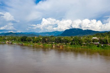 Mekong Nehri, Laos 'un Luang Prabang şehrinde ağaçlarla ve demirli teknelerle dolu yemyeşil bir nehir kıyısını geçer. Uzak mavi dağların üzerinde dramatik kümülüs bulutları yükselir, parlak gün ışığı altında canlı bir tropikal manzara yaratır..