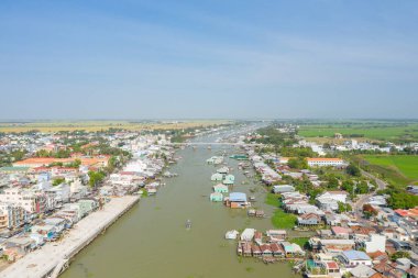 Vietnam, Chau Doc 'un geniş hava manzarası, nehir kenarında evleri olan yüzen bir köy, kıyılar boyunca kentsel binalar ve açık bir gökyüzünün altında uzanan yeşil pirinç tarlaları gösteriyor. Kompozisyon benzersizliği vurguluyor.