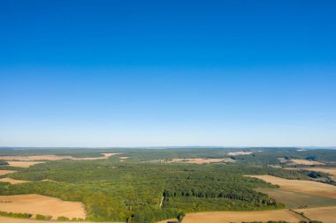 Geniş hava manzarası, Cuncy les Varzy yakınlarındaki kırsal alanda geniş, bulutsuz bir mavi gökyüzünün altında altın tarlalarla çevrili yoğun yeşil ormanlara sahiptir. Açık arazi ve uzak ufuklar bir berraklık ve boşluk hissi uyandırıyor.