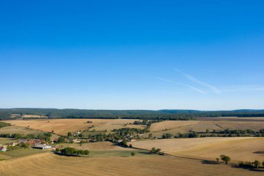 Fransa 'nın orta kesimindeki canlı mavi gökyüzünün altındaki uzak ormanlık tepelerle küçük bir köyü çevreleyen altın tarlalar ve dağınık ağaçların geniş hava manzarası. Açık arazi ve yamalı tarım arazisi bir ferah hissi uyandırıyor.