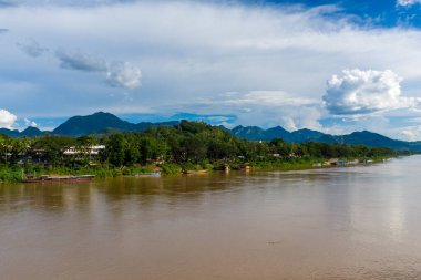 Luang Prabang, Laos 'ta yemyeşil nehir kıyılarından akan Mekong Nehri' nin geniş manzarası ve demirli ahşap kayıklar. Dramatik bulutlar ve uzak dağlar parlak gün ışığı altında sakin tropikal manzarayı çerçeveler..