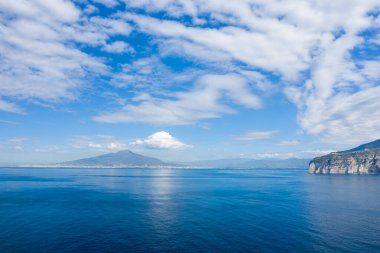 Uzak kıyı şeridinden yükselen Vezüv Dağı 'nın geniş manzarası, Napoli Körfezi' nin sakin mavi suları ve gökyüzünü kaplayan dramatik bulut oluşumları. Sahne, gün ışığı altında ikonik İtalyan kıyılarını yakalıyor..