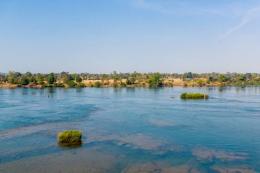 Güney Laos 'un 4000 ada bölgesinde Mekong Nehri' nin geniş bir bölümü küçük yeşil adacıkları ve ağaçlarla kaplı bir nehir kıyısını geçerek akar. Kıtır gün ışığı, durgun mavi suyu ve dağınık bitki örtüsünü vurgular, sakin bir nehir manzarası yaratır..