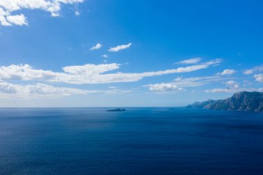 Ufukta küçük adalar ve sağda engebeli kıyı dağları olan engin mavi deniz, dağınık beyaz bulutlarla dolu bir gökyüzünün altında. Sahne aydınlık gün ışığıyla aydınlatılıyor, sakin ve açık atmosferi vurguluyor.
