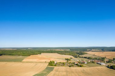 Geniş hava manzarası, altın tarlaların ve ağaçlarla çevrili küçük bir köy ve canlı mavi gökyüzünün altındaki yuvarlanan tepelerin bir kısmını ortaya çıkarıyor. Geniş manzara ve açık ufuk, açık ve kırsal alanda sükunet hissi uyandırıyor.