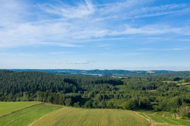 Lac des Settons 'un yoğun kozalaklı ormanları, yamalı tarlaları ve parlak mavi bulutların altında yuvarlanan tepelerle çevrili geniş insansız hava aracı görüntüsü..