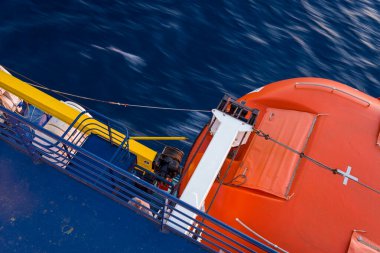 Parlak turuncu bir cankurtaran botu mavi ve sarı bir feribot güvertesinin yanında, aşağıda hareket eden derin mavi bir su var. Görüntü koyu renk karşıtlıkları, geometrik çizgiler ve deniz güvenliği teması içerir..