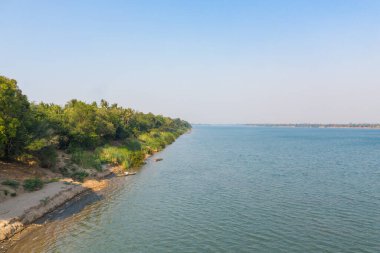 Bereketli bitki örtüsü, Mekong 'un kumlu nehir kıyısında, Stoeng Treng, Kamboçya yakınlarında, berrak mavi bir gökyüzünün altında. Sakin su ve dağınık küçük tekneler Güneydoğu Asya 'da sakin bir nehir kenarı manzarası yaratıyor..