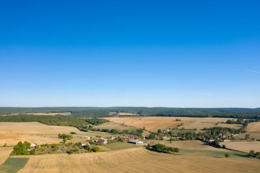Geniş altın tarlalar, dağınık ağaçlar ve canlı mavi gökyüzünün altındaki uzak ormanlık alanlarla çevrili küçük bir kırsal köyün geniş hava manzarası. Açık arazi ve nazik tepeler merkezde bir ferah ve kırsal huzur hissi yaratıyor.