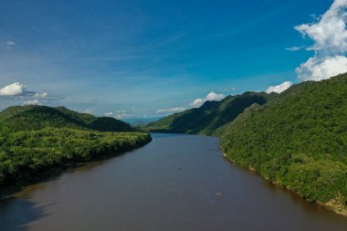 Mekong Nehri 'nin geniş bir bölümü yoğun ormanlık yeşil tepeler arasında dağınık bulutlarla dolu canlı mavi bir gökyüzünün altında akar. Sahnede Güneydoğu Asya 'da zengin tropikal bitki örtüsü, yumuşak yamaçlar ve sakin sular yer alıyor..