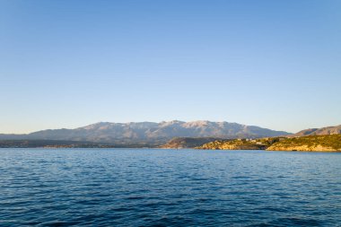 Sıcak akşam güneşi uzak engebeli dağları ve Girit 'in Marathi kenti yakınlarındaki sakin mavi suları aşan kayalık bir kıyı şeridini aydınlatır. Açık gökyüzü ve sakin deniz huzurlu bir Akdeniz manzarası yaratır..
