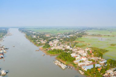Nehir kıyısı boyunca uzanan Chau Doc köyünün geniş hava manzarası, yüzen evler, kümelenmiş evler ve sisli mavi gökyüzünün altında geniş yeşil pirinç tarlaları. Manzara, Mekong 'un kırsal karakterini ve su bazlı yaşam tarzını gözler önüne seriyor.