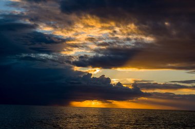 Adriyatik Denizi 'nin yukarısında kara fırtına bulutları toplanırken, parlak altın güneş ışığı su üzerinde çarpıcı ışınlar ve yansımalar yaratıyor. Sahne, gün batımında aydınlık gökyüzü ile derin gölge arasında güçlü bir zıtlık yakalar..