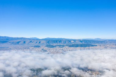 Yunanistan 'ın Epirus kentindeki manzaranın üzerinde yoğun bir alçak bulut tabakası sürüklenen Ioannina şehri ve Pindus dağlarının havadan geniş açılı görüntüsü. Görüntüde canlı gün ışığı, açık mavi gökyüzü ve atmosferik bulut örtüsü var..