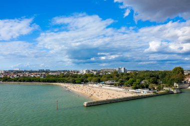 Geniş bir manzara La Rochelle, Charente-Maritime 'in kumlu sahili ve ağaçlarla çevrili kıyı şeridini, modern binalar ve arka planda konut çatıları ile görüntüler. Sahnede turkuaz su, sahile gidenler ve canlı bir gökyüzü yer alıyor.