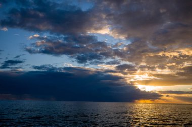 Adriyatik Denizi üzerindeki dramatik bulut oluşumlarından geçen altın güneş ışığı, gün batımında sıcak yansımalarla suyu aydınlatıyor. Geniş deniz manzarası, kara bulutlar ve parlayan gökyüzü arasında çarpıcı bir kontrastı temsil eder..