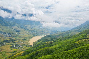 Verimli yeşil tepeler ve katmanlı pirinç terasları Sapa Vadisi, Vietnam 'daki sakin bir dağ gölüne doğru iniyor. Alçak bulutlar arazide sürükleniyor, barışçıl ve atmosferik bir manzara yaratıyor..