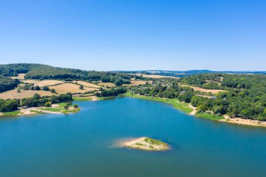 Panneciere-Chaumard rezervuarının hava manzarasında, Burgundy 'de açık bir yaz gökyüzü altında, sakin mavi bir ada, ve orman tepeleriyle çevrili yuvarlanan tarım arazisi görülüyor. Manzara, nazik hatları, yemyeşil ve sakin kırsal kesimleri vurguluyor.