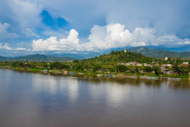 Luang Prabang, Laos 'taki Mekong Nehri' ne bakan yemyeşil bir tepeyi altın bir stupa taçlandırıyor. Renkli tekneler dramatik kümülüs bulutları ve uzak mavi dağların altında nehir kıyısına dizilir..