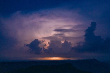 Karanlık fırtına bulutları, uzak bir dağ sırtının üzerinde gökyüzünde parlayan yıldırımlarla parıldıyor. Görüntü dramatik bulut oluşumlarını, katmanlı siluetleri ve karamsar bir alacakaranlık atmosferini görüntüler..