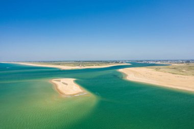 Barre dEtel kumsalının turkuaz suyu ve açık mavi gökyüzünün altında altın kumlu kıyıları olan geniş hava manzarası. Sakin kıyı sahnesinde canlı renkler ve süpürücü doğal formlar yer alıyor..