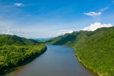Mekong Nehri 'nin açık mavi gökyüzünün altındaki yoğun ormanlık yeşil tepeler arasında kıvrımlı bir görüntüsü. Manzarada canlı tropikal bitki örtüsü, yumuşak yamaçlar ve sakin sular yer alıyor ve Güneydoğu 'da dokunulmamış doğal güzellik hissi uyandırıyor.