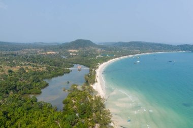 Kamboçya 'daki Koh Rong adasında, beyaz kumla buluşan turkuaz suyu ile Long Set Sahili' nin süpürme yayını gösteren hava görüntüsü. Yoğun yeşil orman, yumuşak dalgalar ve dağınık tekneler sakin bir tropikal kıyı şeridi oluşturur..