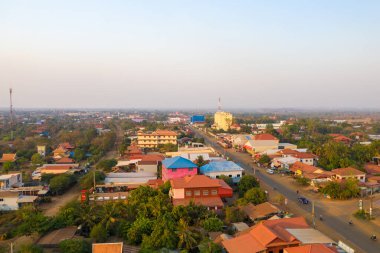 Wide aerial perspective of Krong Preah Vihear city in northern Cambodia, showing colorful rooftops, tree-lined streets, and low-rise buildings under soft evening light. The urban landscape is surrounded by greenery and a hazy horizon.
