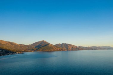 Altın güneş ışığı, İtalya 'nın başkenti Capitello yakınlarındaki sakin mavi bir körfezin üzerinde yükselen yuvarlanan dağları yıkıyor. Açık gökyüzü ve yumuşak su davetkar ve huzurlu bir manzara yaratır..