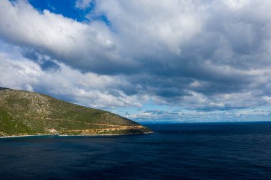 Derin mavi Akdeniz sularına kadar uzanan engebeli yeşil bir yamacın üzerinde dokulu bulutlarla dolu dramatik bir gökyüzü beliriyor. Manzara, Monemvasia yakınlarındaki vahşi, evcilleştirilmemiş kıyı şeridini kayan ışık altında yakalıyor..