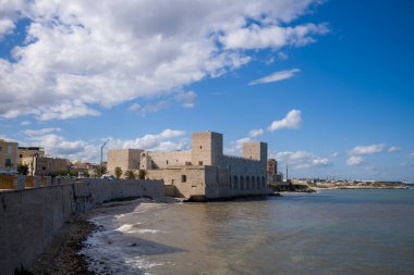 İtalya 'nın Trani kentinde Adriyatik Denizi ile çevrili, kare kuleleri olan sağlam bir ortaçağ taş şatosu bulunuyor. Tarihi kale, gezinti güvertesinde dağınık bulutlar ve palmiye ağaçlarıyla dolu canlı mavi bir gökyüzüne karşı kurulmuştur..