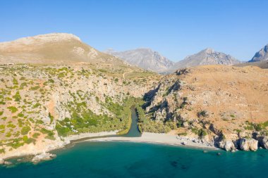 Girit 'in Preveli kentindeki kumlu bir sahil ve turkuaz denizle buluşmak için kayalık uçurumlardan akan palmiye ağaçlarıyla dolu yemyeşil bir nehrin havadan görünüşü. Parlak güneş ışığı, açık mavi gökyüzünün altında yeşil bitki örtüsü ile kurak dağlar arasındaki zıtlığı vurgular..