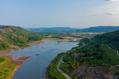 Nehir barajının ve yemyeşil bir tepelik vadideki rezervuarın havadan görüntüsü. Sahnede virajlı yollar, ormanlık yamaçlar ve berrak bir gökyüzünün altında ufku çevreleyen uzak dağlarla birlikte açık su yer alıyor..