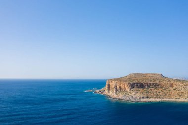 Yunanistan 'ın Girit kentindeki Balos Sahili' nin canlı mavi sularının üzerinde bulutsuz bir gökyüzünün altında güneşli bir kayalık yükseliyor. Sahne açık, sakin ve doğal, sert dokuları ve çarpıcı zıtlıkları var..