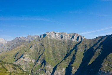 Geniş yeşil yamaçlar ve engebeli kayalıklar Pireneler 'deki Gavarnie Gedre yakınlarındaki bu güneşli dağ manzarasına hakim. Derin gölgeler, canlı mavi gökyüzünün altında dramatik çizgiler çizer..