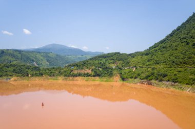 Geniş, sakin, çamurlu bir nehir yemyeşil tepelerin ve Kuzey Vietnam 'daki uzak dağların altında uzanır. Açık gökyüzü ve güneş ışığı suyun üzerinde yumuşak yansımalar ve sıcak toprak renkleri yaratır..