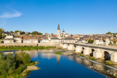 Altın güneş ışığı La Charite sur Loire 'deki tarihi taş köprü ve pastel evleri aydınlatırken, manastır nehir kenti üzerinde yükseliyor. Sakin mavi su ve yemyeşil çerçeve Bu manzaralı Loire Valley manzarası berrak bir gökyüzü altında.