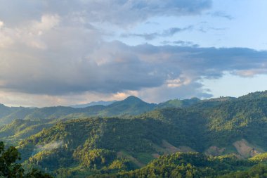 Yumuşak altın güneş ışığı, Vietnam 'da Dien Bien Phu yakınlarında sık ormanlarla kaplı yuvarlanan yeşil dağları vurguluyor. Dramatik bulutlar yumuşak gölgeler bırakır, barışçıl ve geniş bir doğal manzara yaratır..