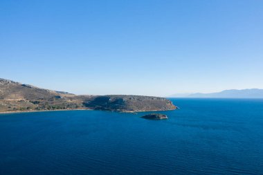 Engebeli bir yarımadanın ve derin mavi sularla çevrili küçük bir adanın geniş bir görüntüsü. Uzak dağlar ve bulutsuz gökyüzü bir açıklık ve sakinlik hissi yaratıyor. Güneşli kıyı şeridinde toprak dokular ve sakin Akdeniz ışığı yer almaktadır..