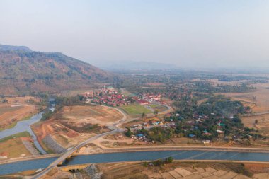 Geniş hava perspektifi, ormanlık tepeler ve puslu gökyüzüne karşı kurulmuş dolambaçlı bir kanal ve yama alanlarının yanında kırmızı çatılı binalar bulunan kırsal bir yerleşim yeri yakalıyor. Manzarada tarım arazisi, dağınık ağaçlar ve uzak bir bölge karışımı yer alıyor.