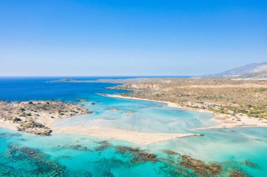 Yunanistan 'ın Girit kentindeki Elafonisi sahilinin geniş çaplı hava görüntüleri, açık mavi bir gökyüzü altında canlı bir turkuaz lagün, kumlu uzantılar ve kurak araziyle çevrili kayalık çıkıntılar gösteriyor. Atmosfer parlak, açık ve doğal olarak davetkar..