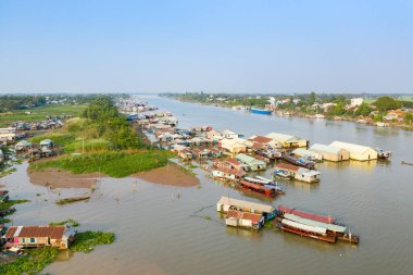 Vietnam 'daki Chau Doc nehri boyunca yüzen bir köyün geniş hava manzarası. Nehir kıyısında demirli evler ve tekneler var. Resimde yemyeşil bankalar, açık kırsal alanlar ve eşsiz nehir kenarını yakalayan açık mavi bir gökyüzü yer alıyor.