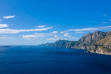 Engebeli kıyı kayalıklarının berrak bir gökyüzünün altında canlı mavi Akdeniz 'le buluşması ufka derinlik katan dağınık bulutlar. Güneş ışığı, İtalyan kıyı şeridinin doğal dokularını ve dramatik hatlarını vurguluyor.